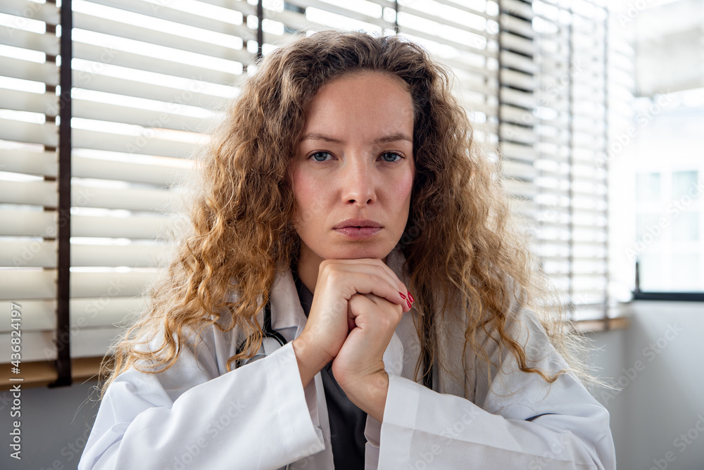 Serious female doctor carefully listening and keeping hands under chin while making medical conference video call from hospital, telehealth concept - obrazy, fototapety, plakaty 