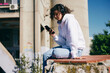 © chika_milan - Cute young woman sitting outdoors with headphones on and choosing good music on her phone.