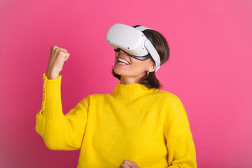 Beautiful woman in bright yellow sweater on pink background in virtual reality glasses happy jumping clenching fist winner gesture