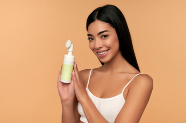 Wall Mural - Young asian woman with radiant skin cleans her face with a brush and foam.isolated on a beige background. Facial skin care concept. Beauty.