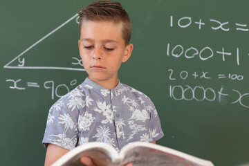 Wall Mural - Caucasian boy standing at chalkboard in classroom holding schoolbook during maths lesson