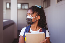 African American Schoolgirl In Face Mask Standing In School Corridor Holding Books