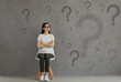 © Studio Romantic - Serious confused woman in glasses sitting on chair looking at multiple question marks on grey background. University, college, high school teen girl doubting, searching, choosing interesting solution