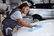 © Roman - Beautiful african woman holds blue microfiber in hand and polishes the car. Cleaning washing auto. Side view portrait of young black auto mechanic woman in uniform at work in car service