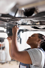 Wall Mural - Black Female Auto Mechanic Working Underneath Car In Garage. Portrait of hardworking young female mechanic in overalls, inspecting lifted car. Mechanic woman working in car repair shop. Side View