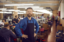 Shoemaker. Portrait Of A Confident Smiling Factory Worker Or Shoe Factory Owner At Work. Caucasian Handsome Bearded Man In A Black Work Apron Holds His Hands To His Sides Looking At The Camera.
