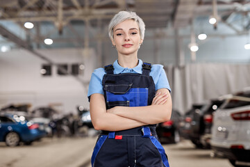 Wall Mural - Caucasian professional female mechanic posing at camera standing in auto repair shop. Short haired woman in blue uniform. Car service, repair, maintenance and people concept. Front view. Copy space
