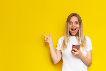 Wall Mural - A young caucasian impressed smiling blonde woman in a white t-shirt points copy empty space for text or design with her finger holding a mobile phone isolated on a bright color yellow background 