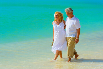 Wall Mural - Older couple hugging and walking on the beach in white outfits 