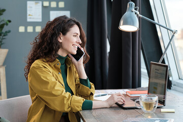 Wall Mural - Young smiling woman sitting at the table working on laptop and talking on mobile phone at home