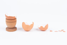 Horizontal Shot Of Broken Egg Shell In Old Wooden Egg Stand And Multiple Other Broken Shells On A White Surface. Minimalism. Conceptual Art For Food Or Animal Products. Sustainability Issue.