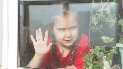 Beautiful little girl smiling and watching out the window. A child looks out the window. Portrait of cheerful kid. self distancing at home during Corona virus. Quarantine Covid-19 World Pandemic.