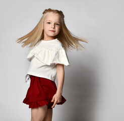 stylish little girl in summer clothes on a white background. Smiling cheerful blonde, dressed in a white blouse and red shorts, turned sharply to the camera, hair flying