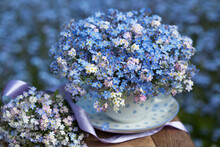 A Bouquet Spring Of Forget-me-nots In A Cup And On A Table On A Blurred Background Of Blue Flowers Outdoor. Blur, Selective Focus, Postcard.