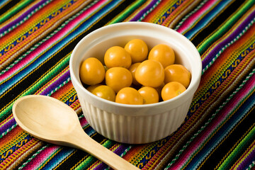 porción de aguaymanto, fruta peruana, en bol blanco y fondo de tela andina de colores con cuchara de madera