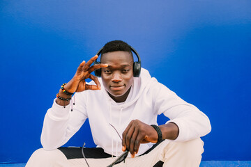 Wall Mural - Young african black man seated against a blue wall looking at camera wearing a white sweatshirt and sunglasses doing the ok sign