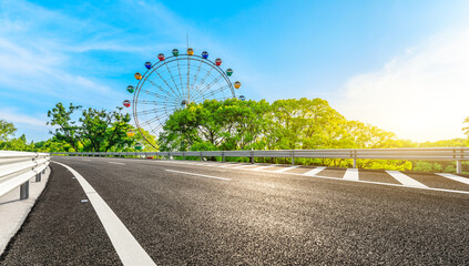 Empty asphalt road and ferris wheel with green forest under blue sky.