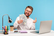 Happy funny man with beard showing fuck gesture with middle finger and dollar cash to camera on laptop talking video call, bragging with job promotion. Indoor studio shot isolated on blue background