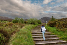Teenager Girl Walking On A Stair Way To Croagh Patrick, County Mayo, Ireland. Cloudy Sky. Mountain Peak And White Statue In The Background. Travel And Adventure Concept