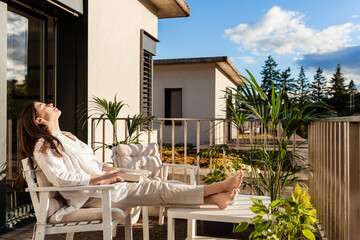 Beautiful young girl with long hair relaxing and enjoying sun sitting at balcony at sunlight at summer.  Slow living, tranquil moment, mental health. Backyard terrace vacation.