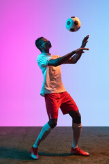 Wall Mural - Portrait of African professional football player standing isolated on gradient blue pink background in neon light.