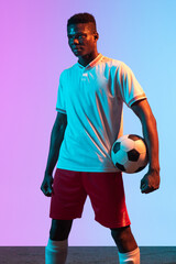 Wall Mural - Half-length portrait of African professional football player standing isolated on gradient blue pink background in neon light.