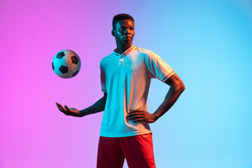 Half-length portrait of African professional football player standing isolated on gradient blue pink background in neon light.