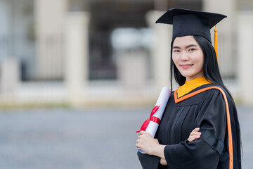 Poster - A young beautiful Asian woman university graduate in graduation gown and mortarboard holds a degree certificate stands in front of the university building after participating in college commencement