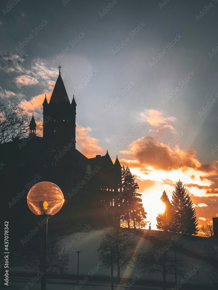 A church stands on a snow-covered hill, silhouetted by the golden rays of the sun setting behind it. - obrazy, fototapety, plakaty 