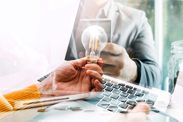 business hand holding lightbulb with using smartphone, laptop computer and money stack in office. idea saving energy and accounting finance concept in morning light