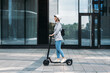 Side view of a young businesswoman riding electric push scooter. Female in cycling helmet driving electrical scooter against building.