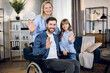 © sofiko14 - Charming mother and cute daughter standing near handsome man that sitting in wheelchair. Happy parents with kid smiling and waving hands at living room.