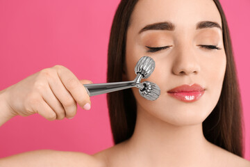 Wall Mural - Woman using metal face roller on pink background, closeup