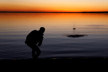 Silhouette Of A Man Throws Stones Into The Lake At Sunset