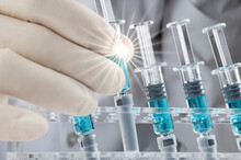 Closed Up Image Of A Doctor Holding A Glass Syringe With Several Other Syringes Prefilled With Blue Liquid On A Rack. Covid-19 Vaccination Concepts.