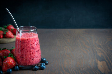 Refreshing sweet smoothie made of fresh juicy strawberries and blueberries served in drinking glass with metal reusable straw on dark brown wooden table against black wall. Image with copy space