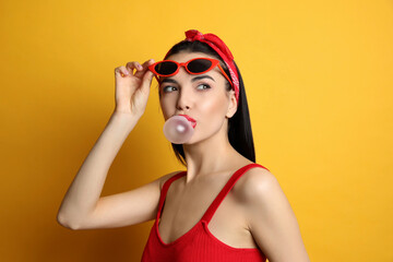 Poster - Fashionable young woman in pin up outfit blowing bubblegum on yellow background