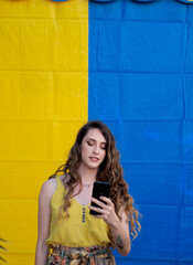 Wall Mural - Vertical closeup shot of a young Caucasian female using her phone near a blue and yellow wall