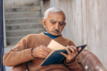 Portrait of old Indian man turning pages of old diary while sitting on chair at home. Leisure activity of retired people. Senior people lifestyle	
