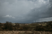 Farm Of Wind Turbines For Renewable Wind Energy On Far Horizon On Hills Of Romania Under The Dark Cloudy Sky. Copy Space.