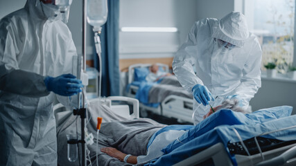 Hospital Coronavirus Emergency Department Ward: Team of Doctors wearing Coveralls, Face Shields Take Care of a Senior Patient Lying in Bed, Put Oxygen Mask and Lung Ventilator. Medics Saving Lives