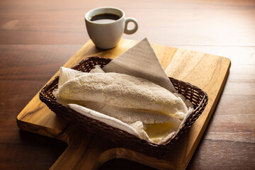 Duas tapiocas dentro de uma pequena cesta de vime e uma xícara de café sobre mesa de madeira.