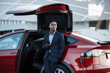 Wall Mural - Confident african man in business suit standing near luxury car with modern smartphone in hands. Handsome male using cell phone while charging battery of his red electric vehicle.