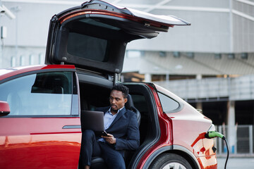 Wall Mural - African american businessman in stylish suit using modern laptop for remote work while his luxury electric car is charging. Handsome male sitting in side auto with opened door and looking aside.