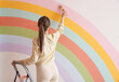 © New Africa - Young woman painting rainbow on white wall indoors, back view. Space for text