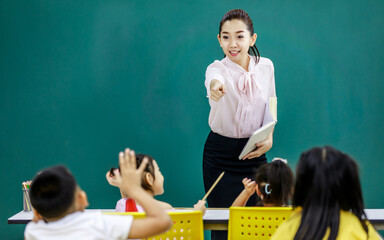 Portrait shot of Asian beautiful female teacher tutor stand smiling holding text book in hands asking question pointing at little clever elementary school boy raising hand up volunteering to answer