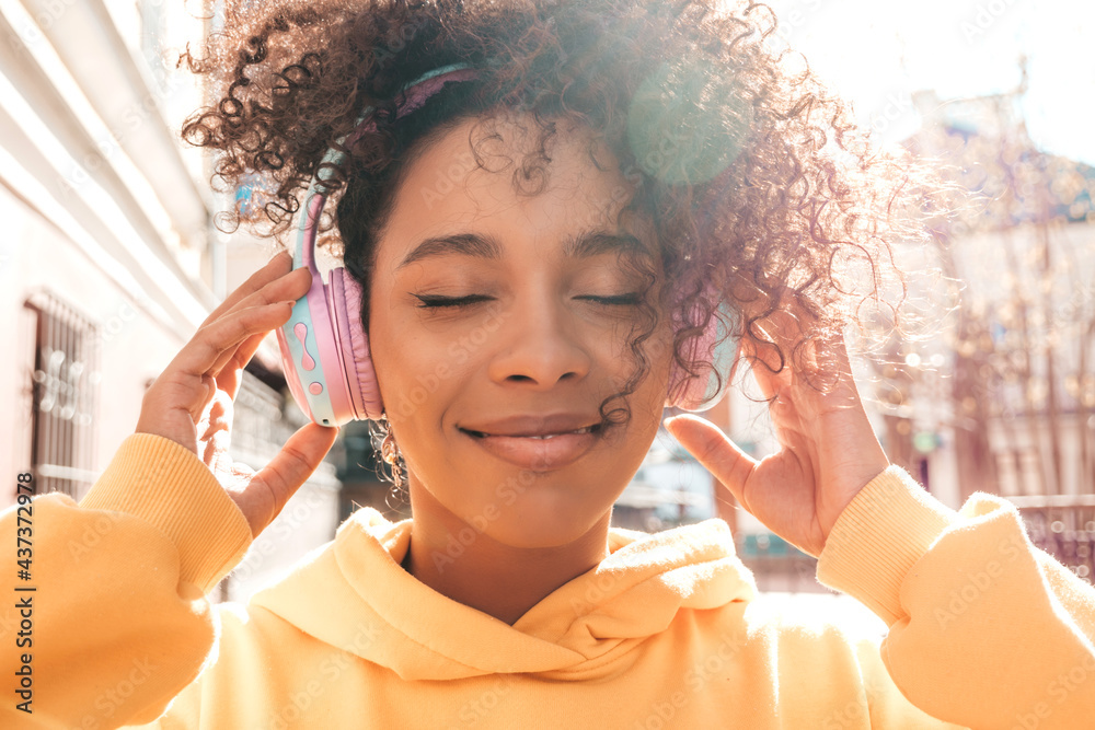 Beautiful black woman with afro curls hairstyle.Smiling model in yellow hoodie.Sexy carefree female enjoying listening music in wireless headphones.Posing on street background at sunset.Closed eyes - obrazy, fototapety, plakaty 