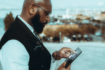 A side view of an elegant mature bald black guy entrepreneur outdoors in eyeglasses and fashionable business outfit, with a well-groomed beard, using a glowing user interface of a futuristic tablet pc
