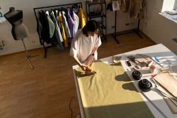 Wall Mural - Sewing process: woman tailor working in shop with chalk patterns on fabric. Top angle view of young clothes designer cut textile for collection in fashion atelier studio. Dressmaking business owner