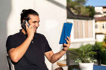 young latino man in a phone call with a note pad in the hand sitting in a balcony